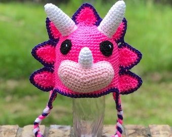 May include: Pink and purple crocheted triceratops hat with white horns and a long, braided strap.