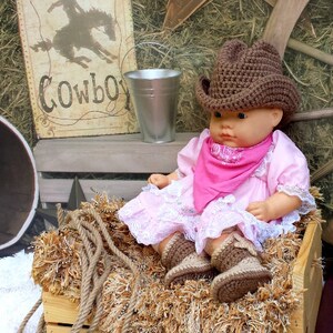 May include: A doll wearing a brown crochet cowboy hat, pink dress, and matching boots, with a pink bandana. The doll is sitting in a wooden crate filled with hay. A sign that says "Cowboy" is in the background.