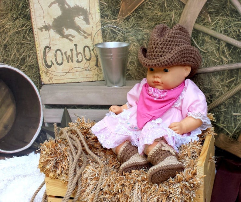 May include: A doll wearing a brown crochet cowboy hat, pink dress, and matching boots, with a pink bandana. The doll is sitting in a wooden crate filled with hay. A sign that says "Cowboy" is in the background.