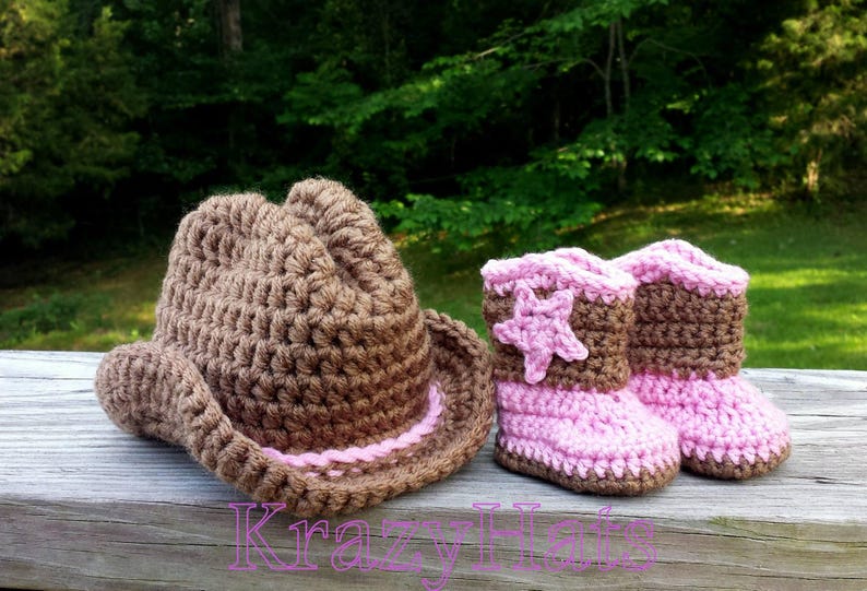 May include: A brown crochet cowboy hat and matching baby boots with pink accents. The boots feature a star design. The items are handmade and suitable for a baby. The word "KrazyHat" is written in pink.