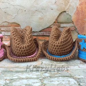 May include: A collection of crocheted cowboy hats and boots in pink, brown, and blue. The boots feature star accents. The hats and boots are arranged in a row against a rustic brick background. The text "KrazyHats" is visible.