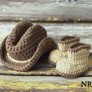 May include: A crocheted brown cowboy hat with a cream-colored band sits next to a pair of matching baby booties. The hat and booties are made of a soft yarn and are displayed on a rustic wooden surface. The text "NR.2" is in the bottom right corner.