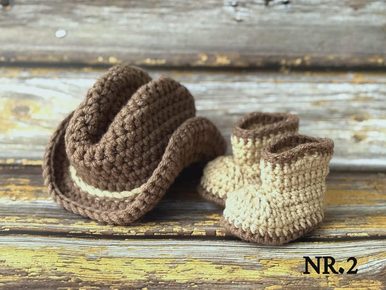 May include: A crocheted brown cowboy hat with a cream-colored band sits next to a pair of matching baby booties. The hat and booties are made of a soft yarn and are displayed on a rustic wooden surface. The text "NR.2" is in the bottom right corner.