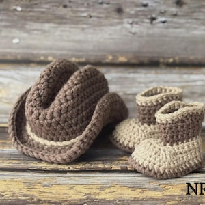 May include: A crocheted baby set featuring a brown cowboy hat and matching booties. The hat has a light tan band, and the booties are two-toned, with a light tan base and brown tops. The set is displayed on a rustic wooden surface.