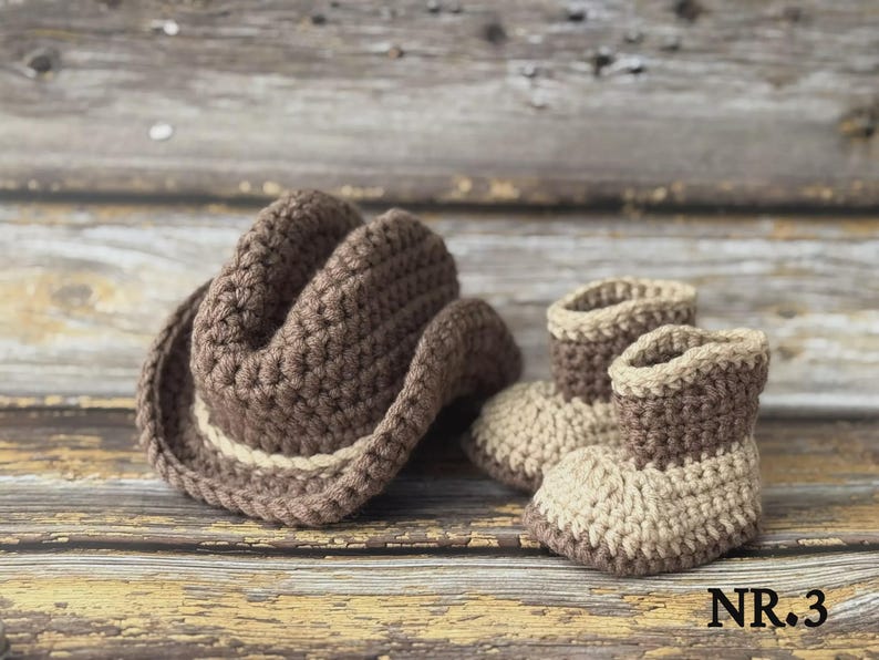 May include: A crocheted baby set featuring a brown cowboy hat and matching booties. The hat has a light tan band, and the booties are two-toned, with a light tan base and brown tops. The set is displayed on a rustic wooden surface.