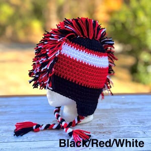May include: A crocheted hat with a mohawk design in black, red, and white. The hat features a striped pattern and braided ties with tassels. The text "Black/Red/White" is at the bottom of the image.
