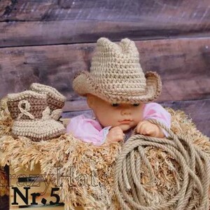 May include: A baby doll wearing a crocheted cowboy hat and boots, resting on a bed of straw. A rope is coiled nearby. The doll is wearing a pink shirt. The background is a wooden wall. The text "Krazy Hats Nr. 5" is visible.