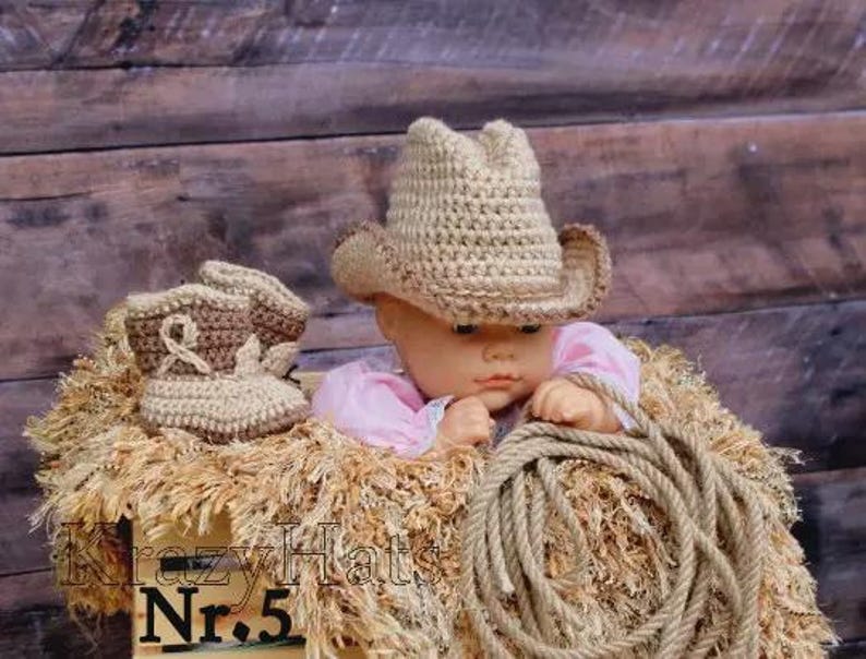 May include: A baby doll wearing a crocheted cowboy hat and boots, resting on a bed of straw. A rope is coiled nearby. The doll is wearing a pink shirt. The background is a wooden wall. The text "Krazy Hats Nr. 5" is visible.
