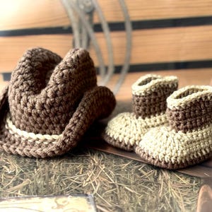 May include: A brown crochet cowboy hat with a cream-colored band sits next to a pair of matching crochet baby cowboy boots. The boots are two-toned, with cream-colored foot sections and brown tops.