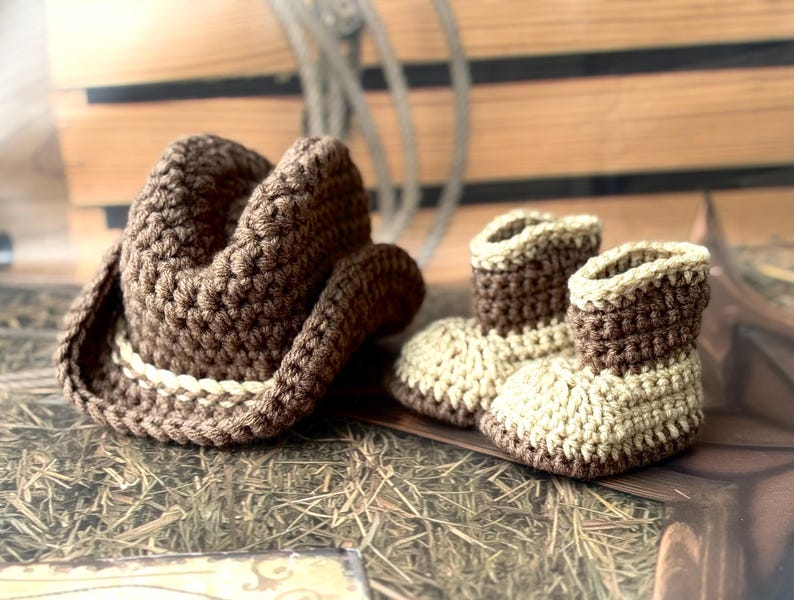May include: A brown crochet cowboy hat with a cream-colored band sits next to a pair of matching crochet baby cowboy boots. The boots are two-toned, with cream-colored foot sections and brown tops.