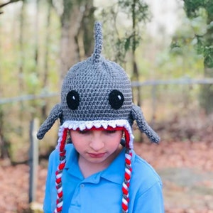 May include: A gray crocheted shark hat with white teeth and red and white braided ties. The hat has black eyes and a gray fin on top.