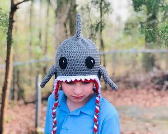 May include: A gray crocheted shark hat with white teeth and red and white braided ties. The hat has black eyes and a gray fin on top.