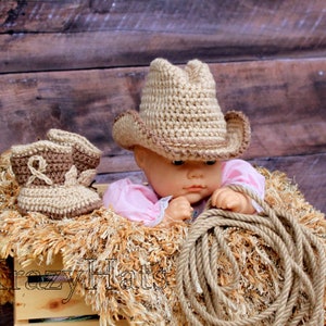 May include: A baby cowboy set featuring a crochet hat and boots in tan and brown. The set is displayed with a doll wearing a pink outfit, a rope, and a wooden crate. The text "Krazy Hats" is visible on the crate.