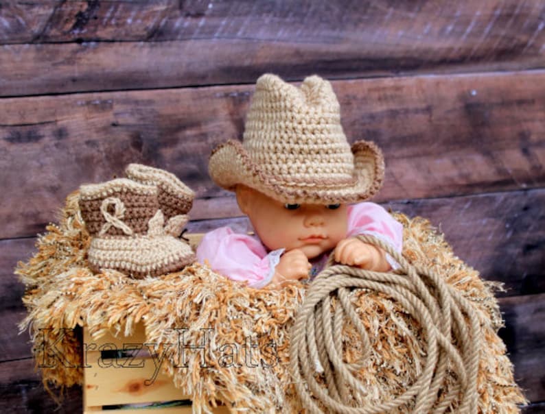 May include: A baby cowboy set featuring a crochet hat and boots in tan and brown. The set is displayed with a doll wearing a pink outfit, a rope, and a wooden crate. The text "Krazy Hats" is visible on the crate.