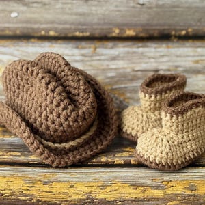 May include: A brown crochet cowboy hat and matching baby boots. The hat has a rounded crown and a wide brim, with a lighter brown band. The boots are tan with brown trim. The set is made of a soft, textured yarn.