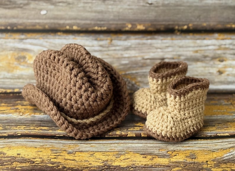 May include: A brown crochet cowboy hat and matching baby boots. The hat has a rounded crown and a wide brim, with a lighter brown band. The boots are tan with brown trim. The set is made of a soft, textured yarn.