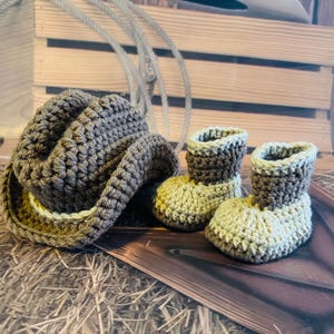 May include: A crochet cowboy hat and two matching baby booties in shades of brown and tan. The hat has a wide brim and the booties are ankle-high. The items are displayed on a wooden surface with a rope in the background.