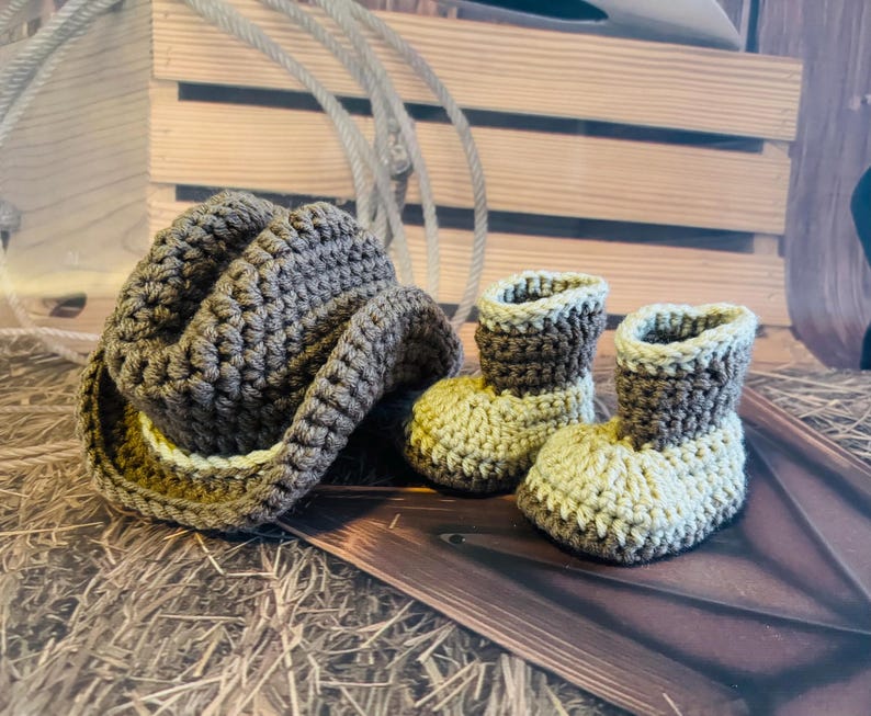 May include: A crochet cowboy hat and two matching baby booties in shades of brown and tan. The hat has a wide brim and the booties are ankle-high. The items are displayed on a wooden surface with a rope in the background.