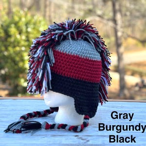 May include: A handmade crochet hat with a mohawk design. The hat features gray, burgundy, and black colors. The hat has ear flaps and braided ties. The text "Gray, Burgundy, Black" is visible on the image.