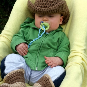 May include: A baby wearing a brown crochet cowboy hat, green hoodie, gray pants, and brown crochet boots, resting in a yellow blanket. The baby has a pacifier in its mouth.