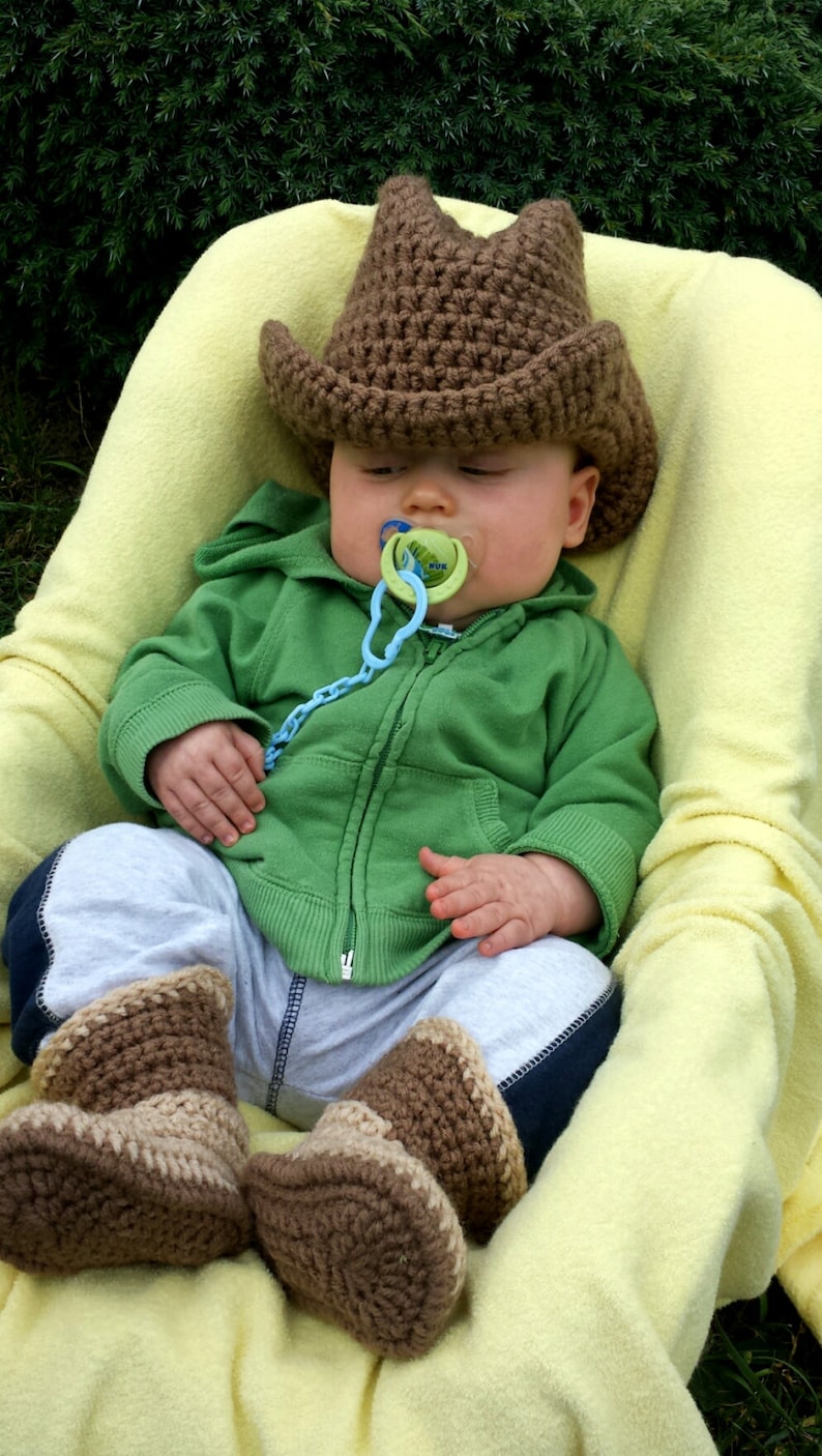 May include: A baby wearing a brown crochet cowboy hat, green hoodie, gray pants, and brown crochet boots, resting in a yellow blanket. The baby has a pacifier in its mouth.