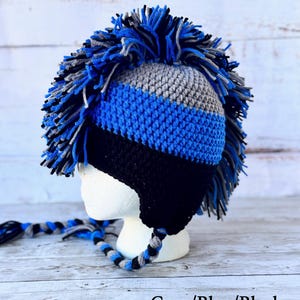 May include: A handmade crochet hat with a mohawk design. The hat features a color block pattern in grey, blue, and black. The mohawk is made of yarn in shades of blue, grey, and black. The hat has ear flaps and a braided tie. The text "Grey/Blue/Black" is at the bottom.