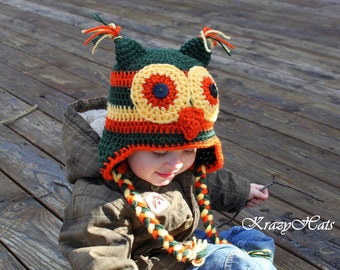 May include: A green, orange, and yellow crocheted owl hat with black button eyes and a long braided cord. The hat is worn by a child sitting on a wooden deck.
