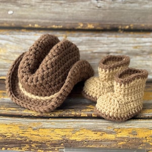 May include: A crocheted cowboy hat and matching baby boots in shades of brown and tan. The hat has a light tan band, and the boots have a brown trim. The set is displayed on a rustic, weathered wooden surface.