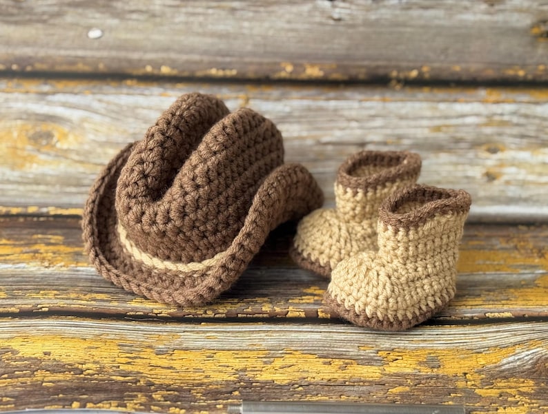 May include: A crocheted cowboy hat and matching baby boots in shades of brown and tan. The hat has a light tan band, and the boots have a brown trim. The set is displayed on a rustic, weathered wooden surface.