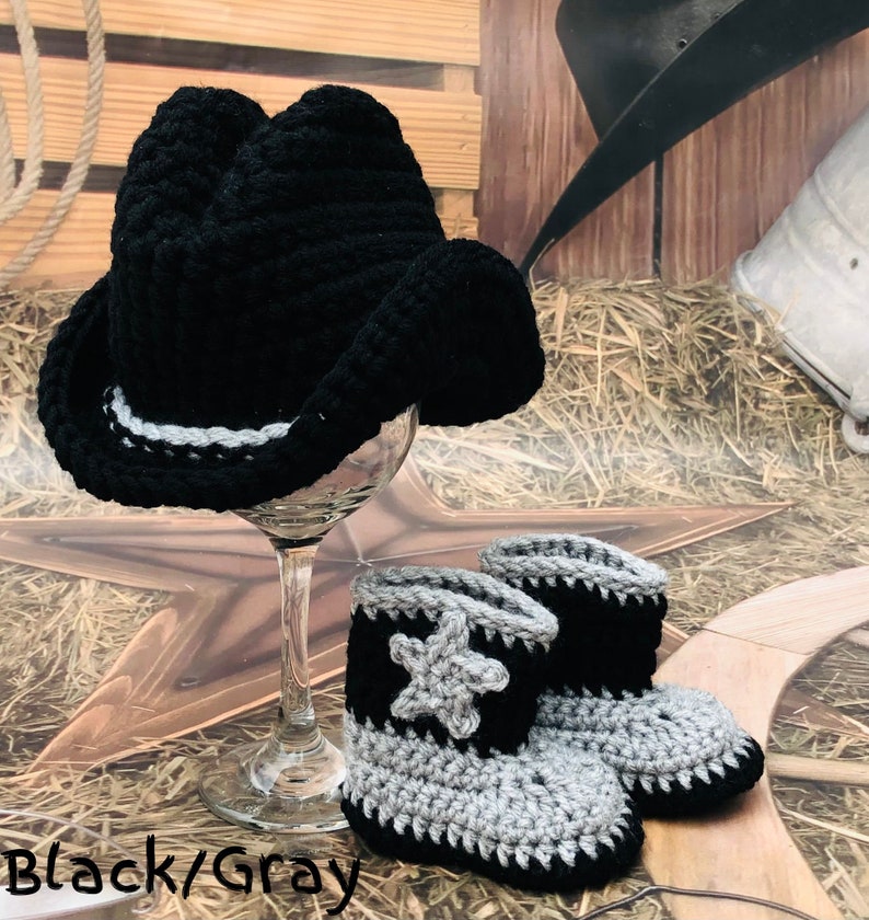 May include: A black crochet cowboy hat with a white and gray band sits atop a wine glass. Matching black and gray crochet baby cowboy boots with a star design are in front of the glass. The text "Black/Gray" is at the bottom.