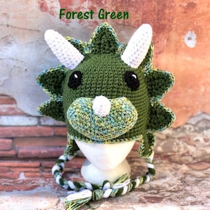 May include: Forest green crocheted triceratops hat with white horns and a white nose. The hat has a long white and green crocheted cord.