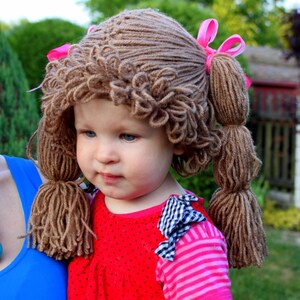 May include: A young child wearing a brown yarn wig with pink bows. The wig has two long pigtails. The child is wearing a red and white polka dot shirt with a pink and white striped sleeve and a black and white checkered bow.