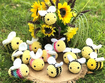 May include: A collection of crocheted bee plushies in various colors, including yellow, black, white, and rainbow. The plushies are arranged on a burlap surface with sunflowers in the background.