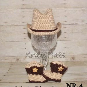May include: A tan crochet cowboy hat with a dark brown band sits atop a glass stand. Below, matching tan and brown crochet cowboy boots with star accents are displayed. The text "Krazy Hats" is visible.
