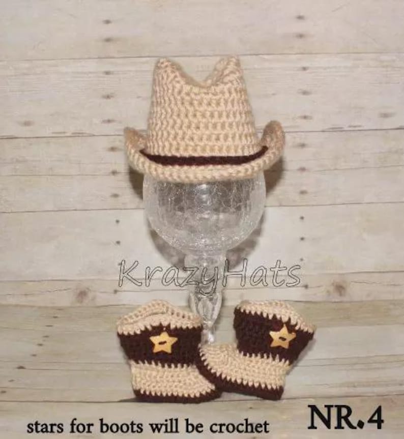 May include: A tan crochet cowboy hat with a dark brown band sits atop a glass stand. Below, matching tan and brown crochet cowboy boots with star accents are displayed. The text "Krazy Hats" is visible.