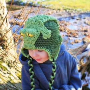 May include: A green crocheted frog hat with black and green braids. The hat is worn by a child with a blue jacket.