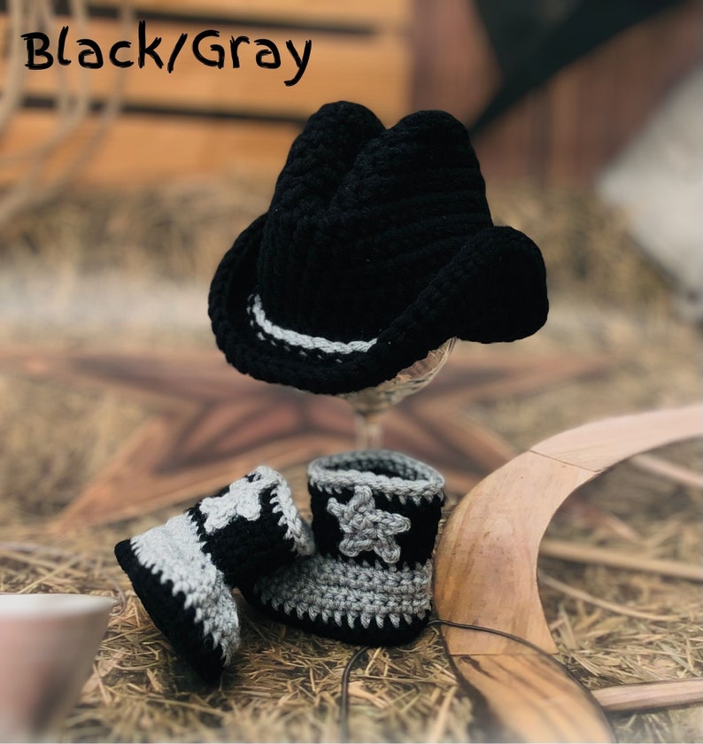 May include: A black and gray crochet cowboy hat and matching baby boots. The hat has a white and gray band. The boots have a star design. The text "Black/Gray" is in the upper left corner.