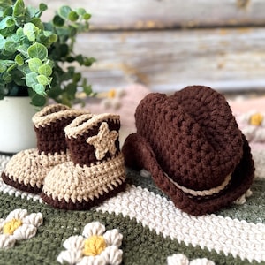 May include: A pair of crochet baby cowboy boots and a matching cowboy hat. The boots are tan and brown with a star applique. The hat is brown with a tan band. The items are on a green and white daisy-patterned blanket.