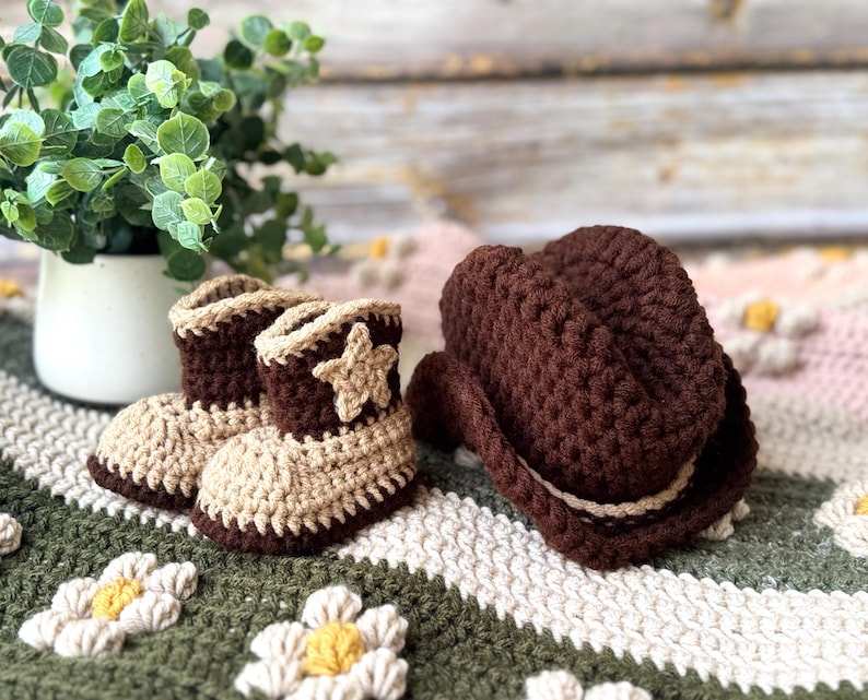 May include: A pair of crochet baby cowboy boots and a matching cowboy hat. The boots are tan and brown with a star applique. The hat is brown with a tan band. The items are on a green and white daisy-patterned blanket.
