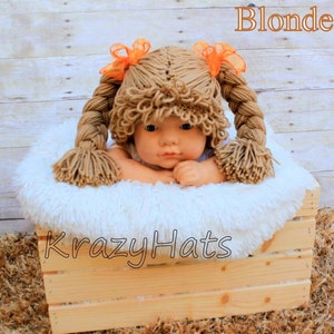 May include: A baby doll wearing a handmade blonde wig with two long braids and orange ribbon accents. The doll is nestled in a white fluffy bed inside a wooden crate. The word "Blonde" is above the doll and "Krazy Hats" is written on the crate.