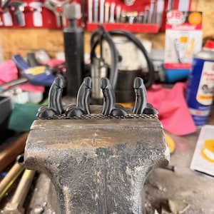 May include: Four small, matte black, phallic-shaped figurines are lined up horizontally and held securely in the metal jaws of a heavy-duty bench vise. The figurines feature a smooth, rounded top and a base with two rounded protrusions on each side.