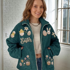 Peut inclure: Une veste zippée en maille gaufrée bleu sarcelle foncé ornée de broderies sur le thème de Pâques, incluant un poussin dans un œuf, un lys, un lapin, une croix, des paniers, des fleurs et le mot "Easter" écrit en lettres blanches.