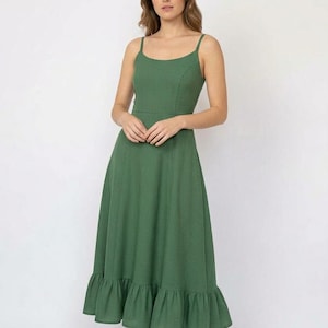 May include: A midi-length olive green dress with a textured waffle-knit fabric, featuring thin spaghetti straps, a fitted bodice, a defined waistline, and a flared skirt that ends in a wide ruffled hem, paired with beige and white athletic trainers.