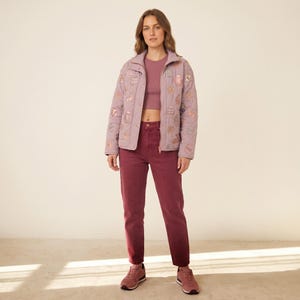 Peut inclure: Une veste matelassée mauve ornée d'un motif répétitif d'œufs de Pâques et de lapins dorés métallisés, portée sur un haut court mauve et un jean rouge foncé. La veste présente un col montant, des manches longues et une fermeture éclair.