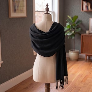 Peut inclure: Une écharpe rectangulaire noire unie drapée sur un mannequin beige. L'écharpe présente une texture douce et mate et est finie avec de longues franges droites le long du bord inférieur, créant une apparence simple et épurée pour l'accessoire.