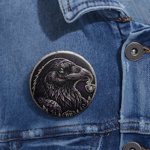 May include: A circular pinback button featuring a detailed illustration of a black raven holding a small steaming cup of coffee in its beak. The background is a dark purple geometric pattern with ornate decorative elements at the bottom.