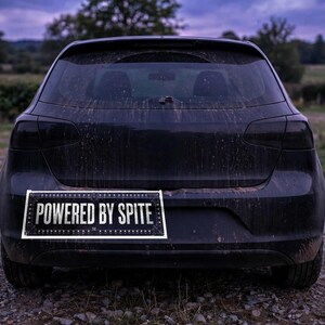 May include: A rectangular bumper sticker with a black background and white text that reads POWERED BY SPITE, featuring a decorative border with small white dots, applied to the rear bumper of a dark, mud-splattered hatchback vehicle.