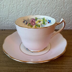 May include: A pale pink porcelain teacup and saucer set featuring a scalloped edge and gold trim. The interior of the cup displays a floral pattern with yellow, purple, and blue flowers. The handle is also accented with gold detailing.