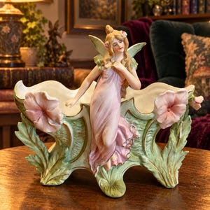May include: A ceramic planter featuring a central fairy figure with long blonde hair wearing a flowing pink dress. The vessel is decorated with sculpted green leaves and large pink flowers on both sides, accented with gold trim along the edges.