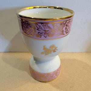 May include: A white porcelain egg cup featuring a wide band of soft pink around the top and base, decorated with intricate gold floral and lattice patterns. The rim is finished with a gold edge, and a small gold floral motif sits in the center.